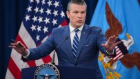 Pete Hegseth speaking passionately at a podium, wearing a suit, with an American flag backdrop and engaging audience.
