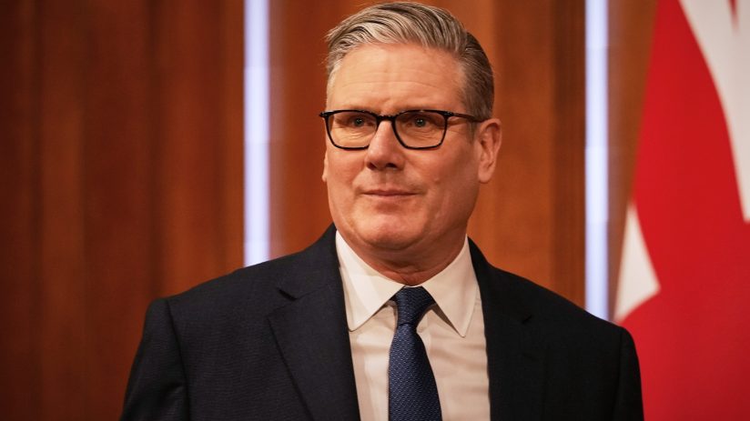 Sir Keir Starmer speaking at a press conference podium, addressing media attendees, wearing a suit and tie, in a formal se...