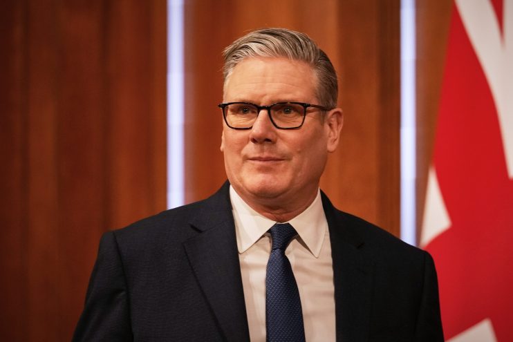 Sir Keir Starmer speaking at a press conference podium, addressing media attendees, wearing a suit and tie, in a formal se...