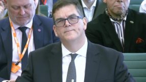 Sir Olly Robbins in a formal setting, possibly during a business or political event, wearing a suit and tie, engaging in d...