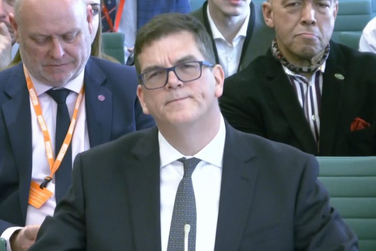 Sir Olly Robbins in a formal setting, possibly during a business or political event, wearing a suit and tie, engaging in d...