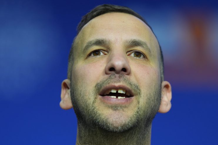 Zack Polanski speaking at a podium during a press conference, wearing a suit and tie, addressing environmental policies