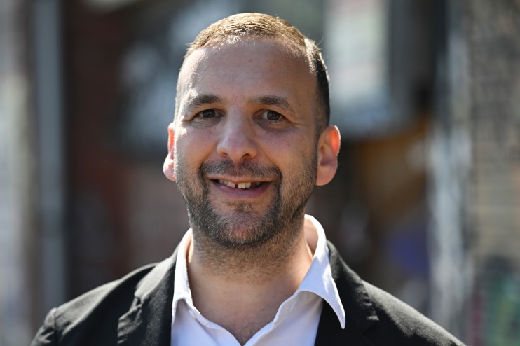 Zack Polanski speaking at a podium during a press conference, wearing a suit and tie, addressing the audience.
