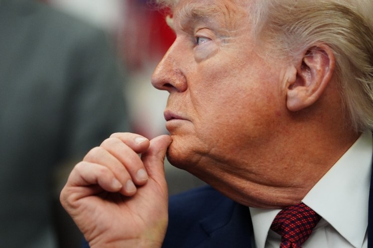 Donald Trump with hand on chin, appearing contemplative during a public event, wearing a suit and red tie.