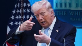 Donald Trump speaking at press conference podium, addressing media with serious expression, American flags in background