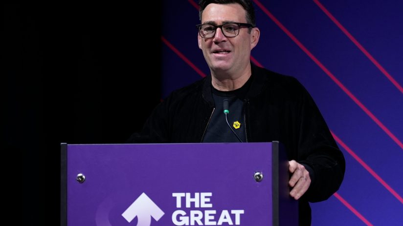 Andy Burnham delivers speech, standing at podium, emphasizing key points, wearing a suit, addressing an engaged audience
