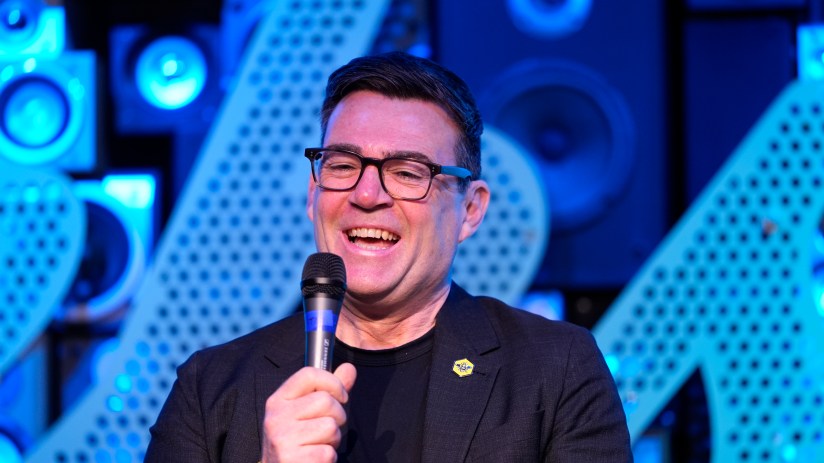 Andy Burnham speaking at a public event, addressing key issues in Manchester, wearing a suit and gesturing with his hands