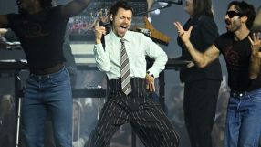 Harry Styles performing on stage with vibrant lighting at a concert, wearing a stylish outfit and engaging with the audience.