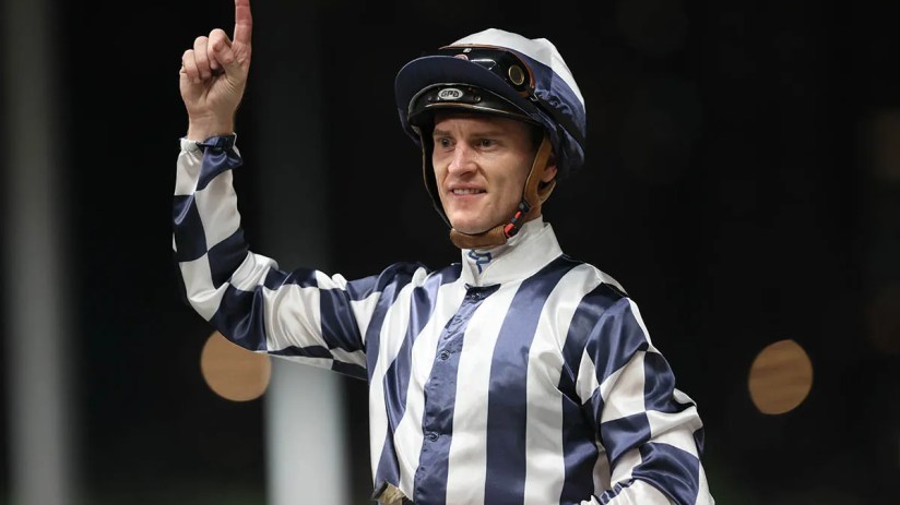 Jockey Zac Purton celebrates after winning a major horse race, wearing his racing silks and holding a trophy aloft.