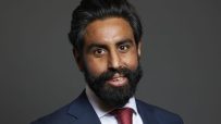 Jeevun Sandher speaking at a business conference podium, addressing the audience with a presentation screen in the backgro...