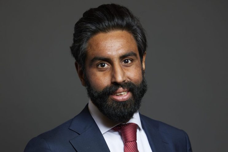 Jeevun Sandher speaking at a business conference podium, addressing the audience with a presentation screen in the backgro...