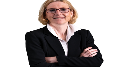 Christiane Ohlgart, a blonde woman in glasses, smiles with arms crossed, wearing a black suit and white shirt.