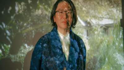 Person with long dark hair and glasses wearing a blue patterned jacket, projection of greenery behind them.