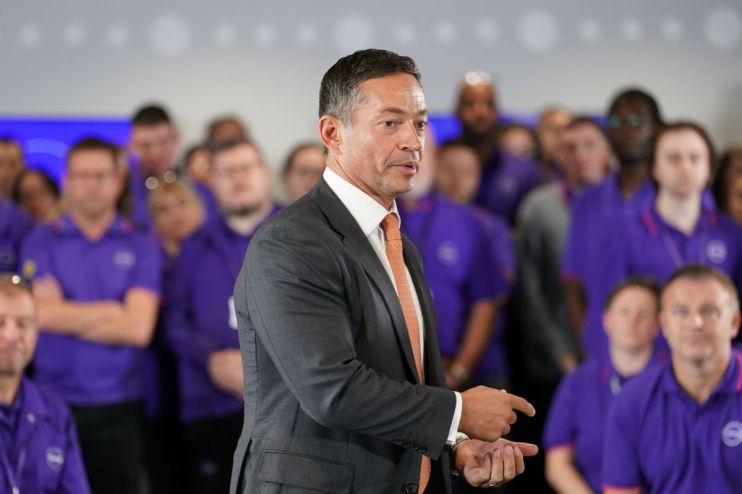 Alex Baldock in a suit and orange tie speaking to a crowd of people in purple shirts.