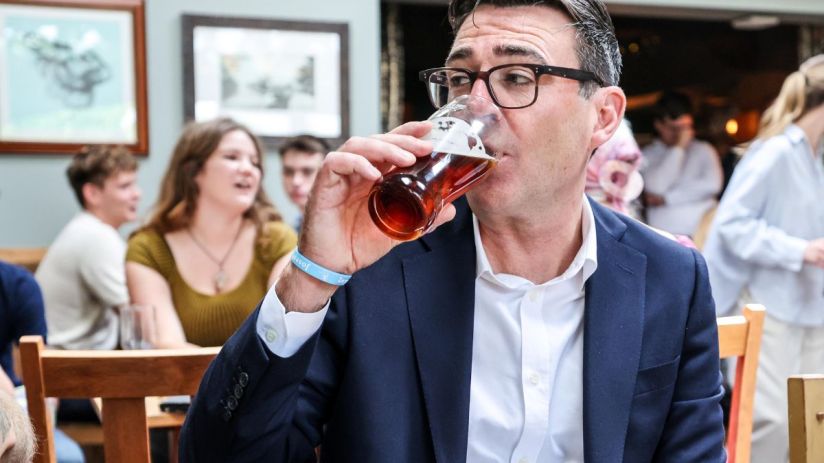 Andy Burnham, Mayor of Greater Manchester, drinks a pint of ale in a pub with people blurred in the background