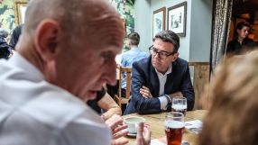 Andy Burnham, Mayor of Greater Manchester, in a dark suit and glasses, listening intently at a wooden table.