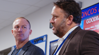 Two businessmen, one with a beard, in a meeting room with a red and blue digital display in the background.