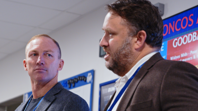Two businessmen, one with a beard, in a meeting room with a red and blue digital display in the background.