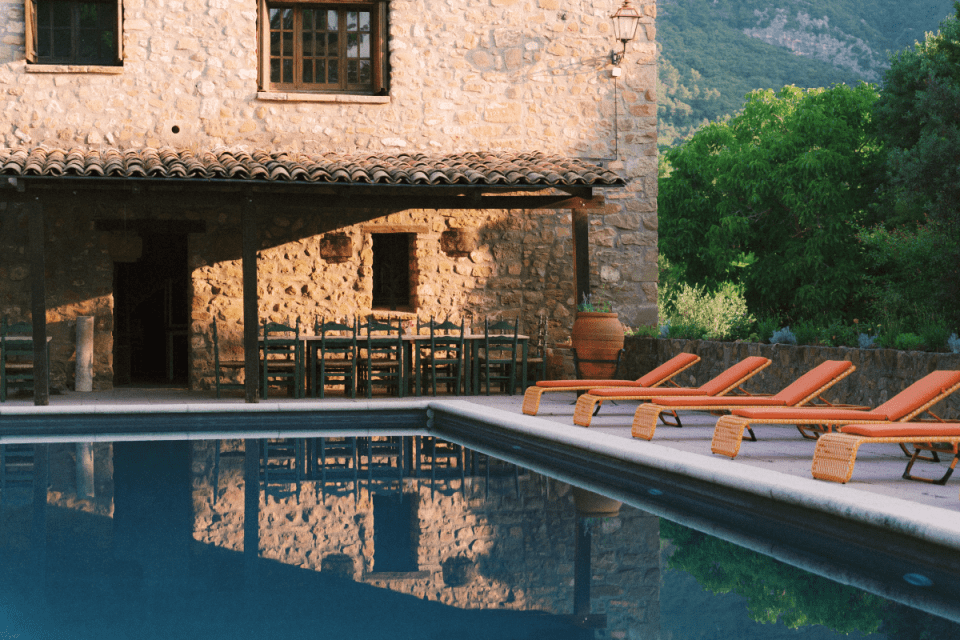 Stone house with covered patio and long dining table next to a swimming pool with orange lounge chairs in Girona.