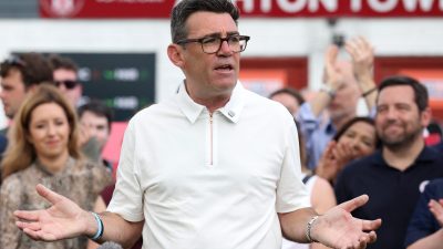 Andy Burnham speaking at a podium with microphones, gesturing with his hands, crowd in background