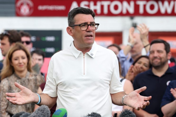 Andy Burnham speaking at a podium with microphones, gesturing with his hands, crowd in background