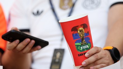 Person holding a red FIFA World Cup branded cup and a smartphone, wearing a white shirt and watch