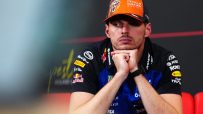Max Verstappen in a Red Bull Racing cap and team shirt, looking contemplative with hands under his chin.