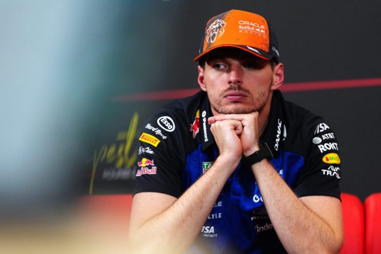 Max Verstappen in a Red Bull Racing cap and team shirt, looking contemplative with hands under his chin.