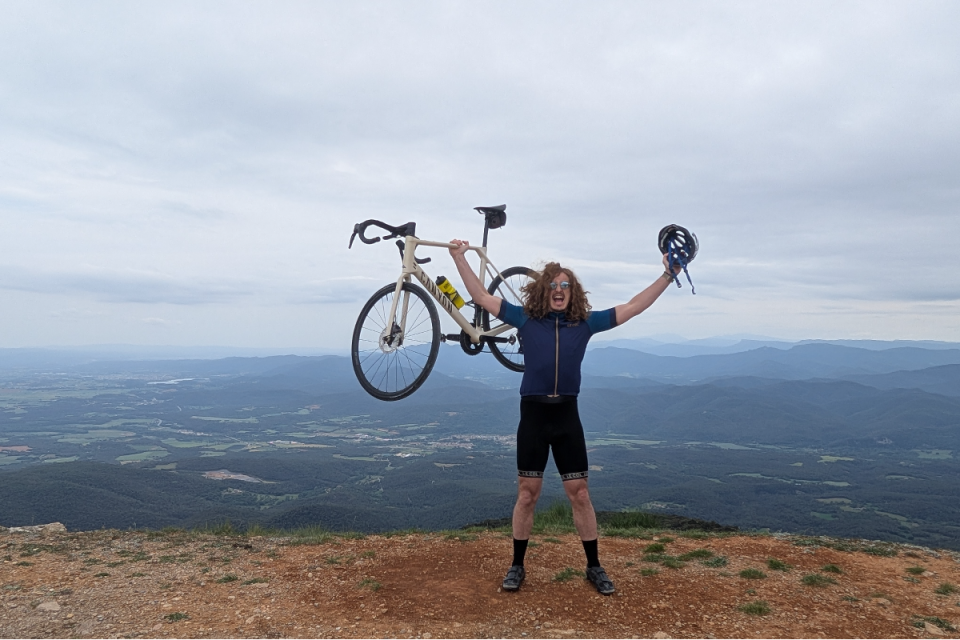 Excited cyclist holding up their bike and helmet on a mountain peak overlooking the Girona landscape.