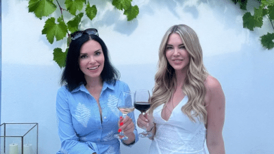 Two women smiling, one in blue holding white Greek wine, another in white holding red wine, under grapevines.