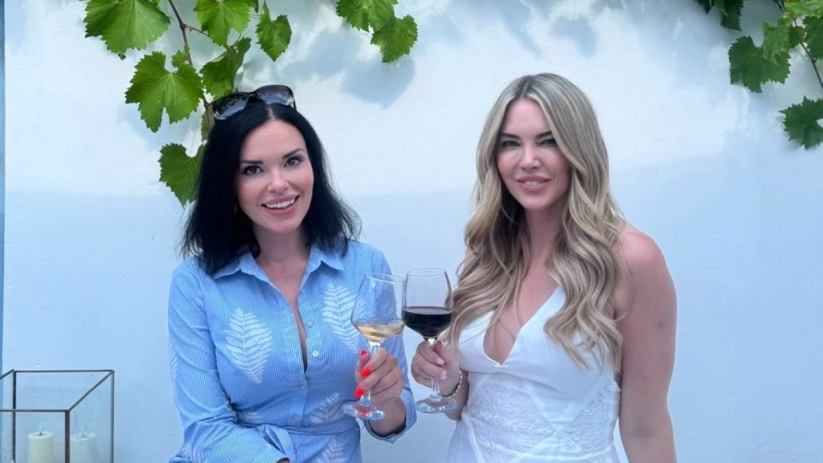 Two women smiling, one in blue holding white Greek wine, another in white holding red wine, under grapevines.