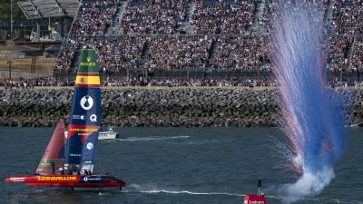 SailGP F50 catamaran Los Gallos sailing past a cheering crowd as red and blue smoke erupts.