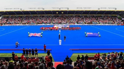 Teams and flags on a blue hockey pitch before a match, with a stadium full of spectators.