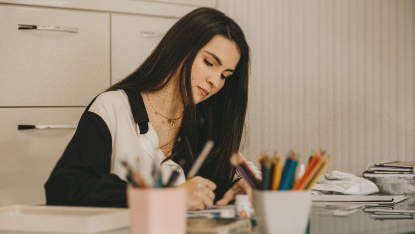 Young woman drawing or writing at a desk, surrounded by art supplies, illustrating human creativity vs AI.