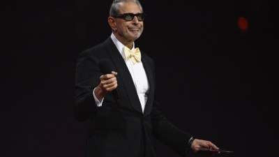 Jeff Goldblum in a black tuxedo with a yellow bow tie and dark sunglasses, holding a microphone, smiling.