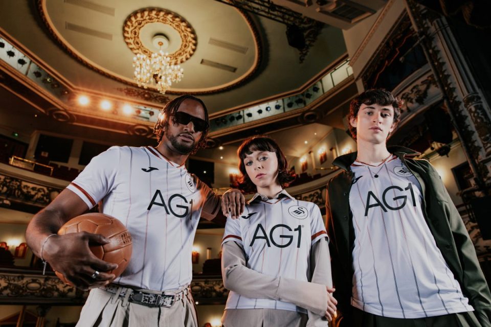 Three models in the new Swansea home kit, featuring vertical stripes, in a grand theater with a chandelier.