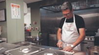 Man in The Passage apron slicing food in a commercial kitchen, with a food allergens chart visible.