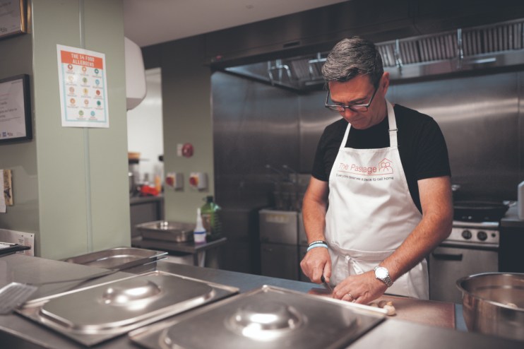 Man in The Passage apron slicing food in a commercial kitchen, with a food allergens chart visible.
