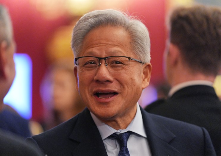 Smiling man with gray hair and glasses in a dark suit and blue tie, speaking at an event.