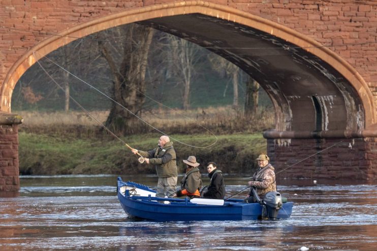 Four people fishing from a blue boat under a brick bridge on a river.
