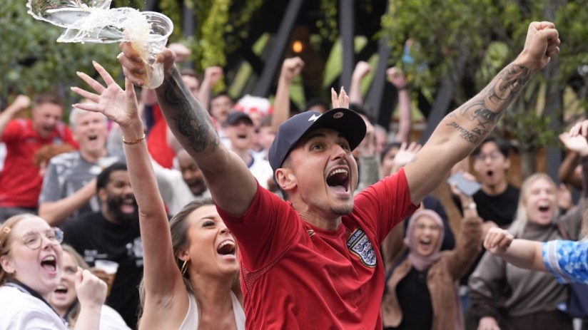 Excited crowd celebrating, a man in an England jersey cheers with arms raised and beer splashing from a cup.