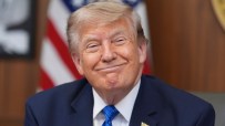 Donald Trump smiling in a blue suit and tie with an American flag pin, US flag in background