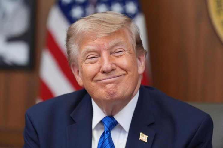 Donald Trump smiling in a blue suit and tie with an American flag pin, US flag in background