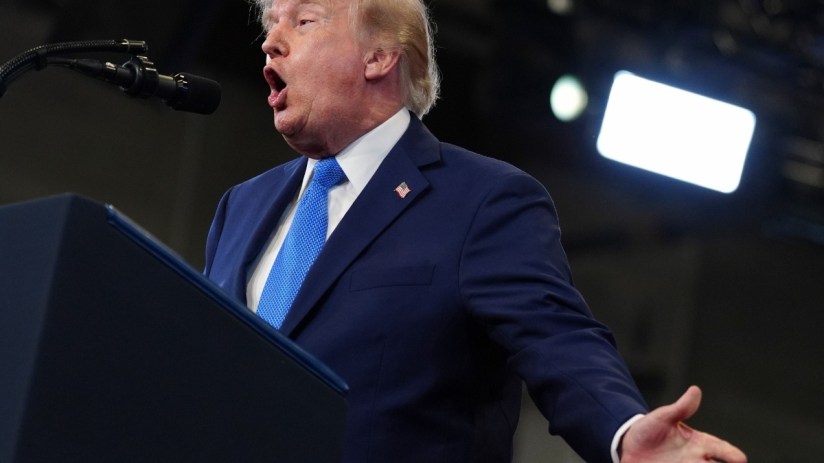 Donald Trump speaking emphatically at a podium, wearing a navy suit and blue tie, with a microphone and lights visible.