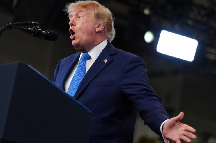 Donald Trump speaking emphatically at a podium, wearing a navy suit and blue tie, with a microphone and lights visible.