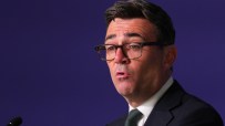 Andy Burnham, Mayor of Greater Manchester, speaking at a podium against a dark blue background.