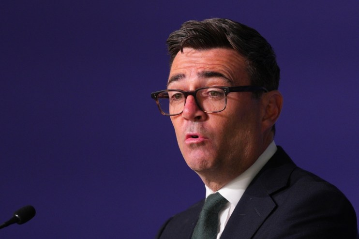 Andy Burnham, Mayor of Greater Manchester, speaking at a podium against a dark blue background.
