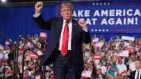 Donald Trump raises a fist to a crowd holding American flags and MAGA signs, with a Make America Great Again! banner behin...
