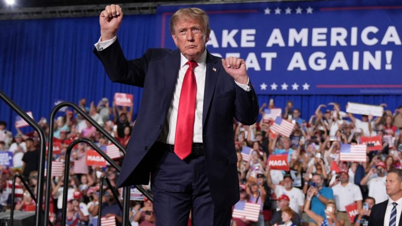 Donald Trump raises a fist to a crowd holding American flags and MAGA signs, with a Make America Great Again! banner behin...
