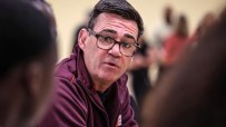 Man in glasses and maroon jacket speaking, with out-of-focus figures in the background.